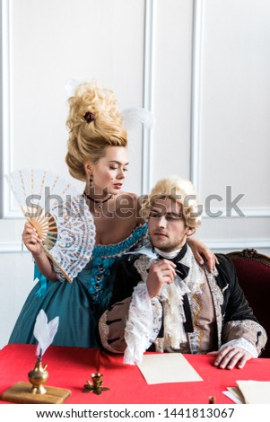 beautiful victorian woman holding fan and standing near pompous man holding feather near papers  Royalty-Free Stock Photo #1441813067