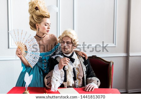 young victorian woman holding fan and standing near pompous man holding feather near paper Royalty-Free Stock Photo #1441813070