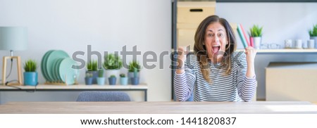 Wide angle photo of middle age senior woman sitting at the table at home celebrating surprised and amazed for success with arms raised and open eyes. Winner concept.