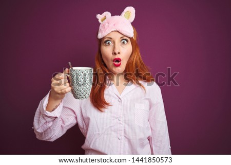Young redhead woman wearing pajama drinking coffee over purple isolated background scared in shock with a surprise face, afraid and excited with fear expression