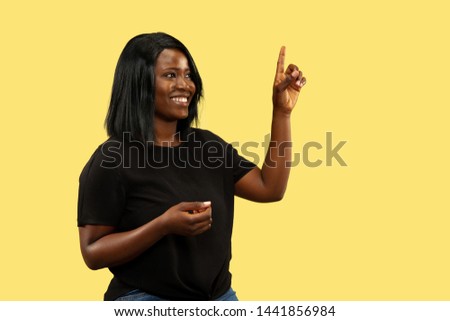 Young african-american woman isolated on yellow studio background, facial expression. Beautiful female half-length portrait. Concept of human emotions, facial expression. Pointin up, touching empty