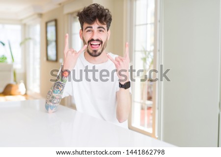 Young man wearing casual t-shirt sitting on white table shouting with crazy expression doing rock symbol with hands up. Music star. Heavy concept.