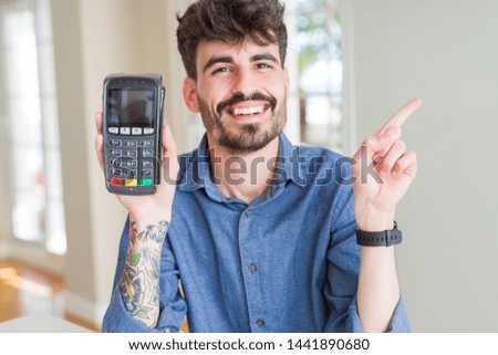 Young man holding dataphone point of sale as payment very happy pointing with hand and finger to the side