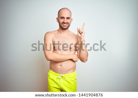 Young shirtless man on vacation wearing yellow swimwear over isolated background with a big smile on face, pointing with hand and finger to the side looking at the camera.