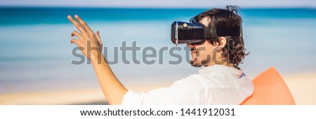 BANNER, LONG FORMAT Summer lifestyle portrait of man sitting on the orange inflatable sofa and uses virtual reality headset on the beach of tropical island. Relaxing and enjoying life on air bed