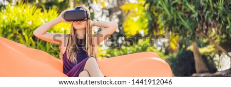 BANNER, LONG FORMAT Summer lifestyle portrait of pretty girl sitting on the orange inflatable sofa and uses virtual reality headset on the beach of tropical island. Relaxing and enjoying life on air