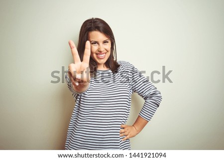 Young beautiful brunette woman wearing striped sweater over white isolated background smiling with happy face winking at the camera doing victory sign. Number two.