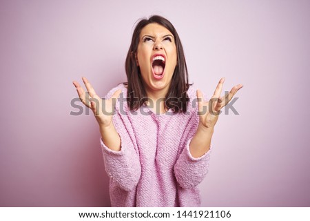 Young beautiful brunette woman wearing a sweater over pink isolated background crazy and mad shouting and yelling with aggressive expression and arms raised. Frustration concept. Royalty-Free Stock Photo #1441921106
