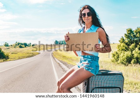 Pretty young woman hitchhiking along a road and waiting on a country road with her suitcase, empty cardboard plate. Beautiful girl in sunglasses hitchhiker. empty Board