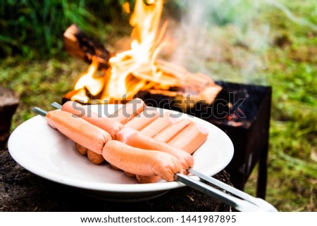 fry sausages on fire. Picnic in nature. Sausages on skewers. Meat products on background of fire with smoke. Eco picnic with reusable dishes. Snack on nature. Grilled sausages.