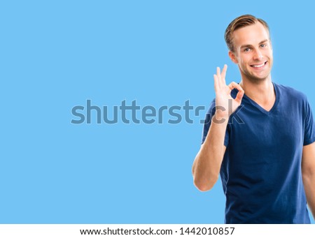 Portrait photo - young happy smiling man in blue casual clothing, showing okay gesture, or zero hand sign, blue color background. Brunette excited male model at studio.