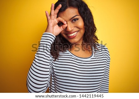 Transsexual transgender woman wearing striped t-shirt over isolated yellow background with happy face smiling doing ok sign with hand on eye looking through fingers