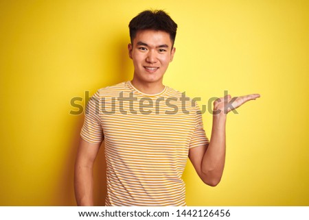 Young asian chinese man wearing t-shirt standing over isolated yellow background smiling cheerful presenting and pointing with palm of hand looking at the camera.