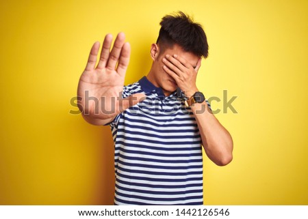 Young asian chinese man wearing striped polo standing over isolated yellow background covering eyes with hands and doing stop gesture with sad and fear expression. Embarrassed and negative concept.