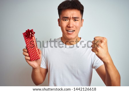 Young asian chinese man holding valentine present standing over isolated white background annoyed and frustrated shouting with anger, crazy and yelling with raised hand, anger concept