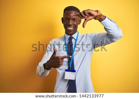 African american businessman wearing identification card over isolated yellow background smiling making frame with hands and fingers with happy face. Creativity and photography concept.