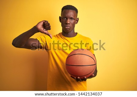 African american athlete man holding basketball ball standing over isolated yellow background with angry face, negative sign showing dislike with thumbs down, rejection concept