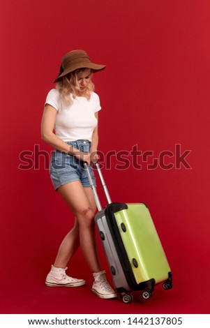 Young blond curly woman in a sundown hat feels upset and tired holding heavy grey luggage bag while standing infront of a red background. Concept of traveling