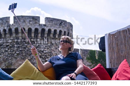 Woman tourist take a selfie photo using smart phone on selfie stick.Sitting on colorful pillows near old stone fortress tower in Belgrade,Serbia