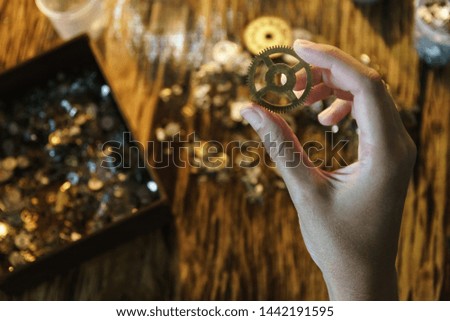 Watchmaker holds large gear. Cogwheel on blurred background of workplace. Pinion in female hand. Old cogwheel on wooden table background. Big metal gearwheel on blurred workspace.