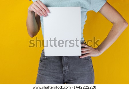 Woman holding white empty sheet of paper for copy space on yellow background. Crop photo, studio shot