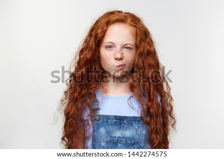 Photo of malcontent nice freckles little girl with ginger hair, parents forbid to watch cartoons, grumpy looks at thecamera, stands over white background.