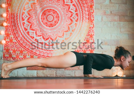 Pretty girl in black body suit doing chaturanga or plank pose in front of a mandala tapestry while yoga practice