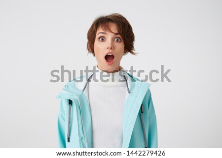 Close up of wondered short-haired young woman in white golf and blue rain coat, stands over white background with wide open mouth and surprised expression, looks at the camera with wide open eyes.