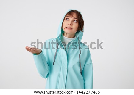 Cute frowning short-haired curly woman in blue rain coat, hiding under the hood from rain and looking up at the rigt side, putting the palm under the rain standing over white background.