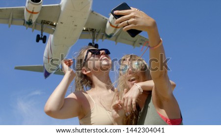 Two young joyful cheerful girls taking a selfie near airport with huge airplane flying over their heads.Best friends taking photos against sky background.Tourism,destination and friendship concept