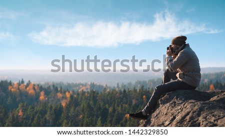 A photographer takes pictures of the landscape. He sits on a stone hill