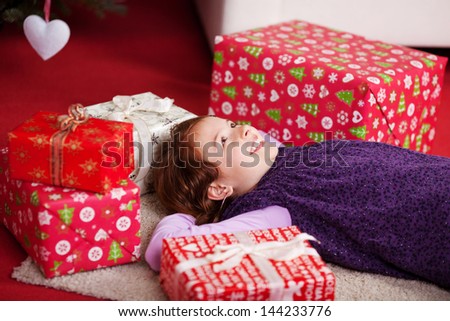 Little girl lying on her back on the living room carpet surrounded by colorful red giftwrapped boxes dreaming of Christmas day
