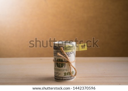 A sticky note and a roll of American currency (USD, American dollars) with 100 dollars bank notes on the outside as a symbol of plenty of money on the wooden background. 