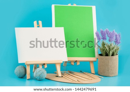 Two wooden blank easels with brushes and plant on blue background.