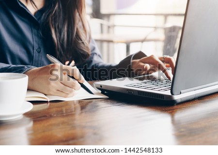 Business woman using laptop and writing on a notepad with a pen at a coffee shop. Working on project concept. Royalty-Free Stock Photo #1442548133