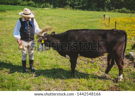 An elderly farmer with a jug in his hand and a hat on his head is standing next to a cow in a pasture.