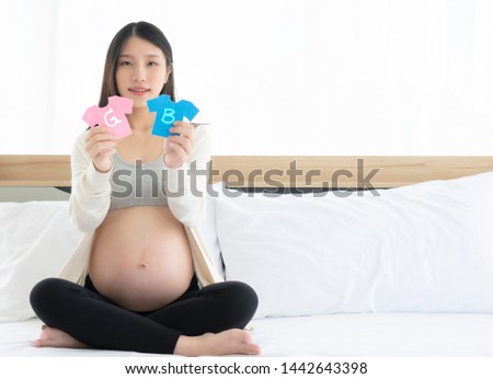 A beautiful Asian pregnant woman sitting on a bed in both hands, showing a small pink and blue T-shirt with a letter G and B in the middle. The concept is the symbol of a Girl and Boy.