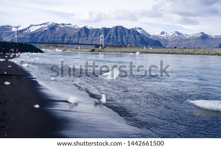 Iceland summer - Jokulsarlon Diamond Beach. Icebergs flowing into Atlantic, some washed up on the black sand beach. Picture taken from the sea towards the lagoon and inland mountains. Bridge and cars 