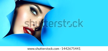 The face of a young beautiful girl with bright makeup and plump red lips looks into the hole in blue paper. Fingers with natural color varnish.