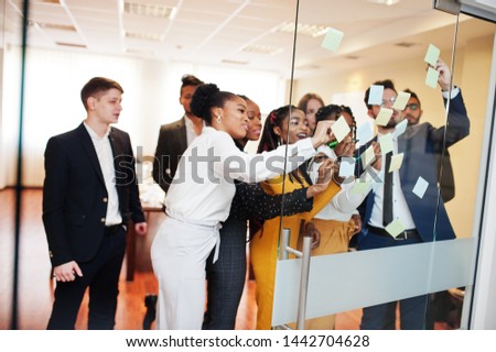 Team of multicultural young people pointing on glass with colorful paper notes. Diverse group of male and female employees in formal wear using stickers. 