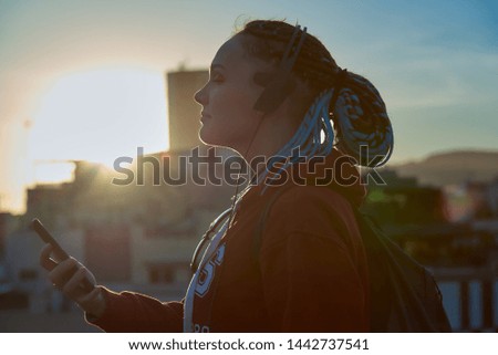 Silhouette of a girl with blue dreadlocks in headphones and with a phone on the background of the sunset.