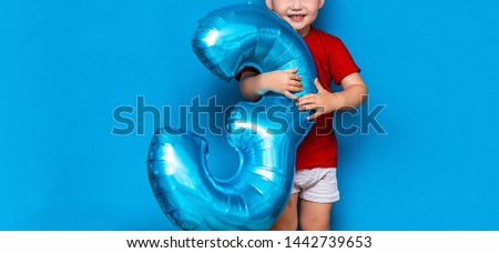 small cute blonde boy on blue background holding foil-coated sphere baloon blue colour. happy birthday three years old.