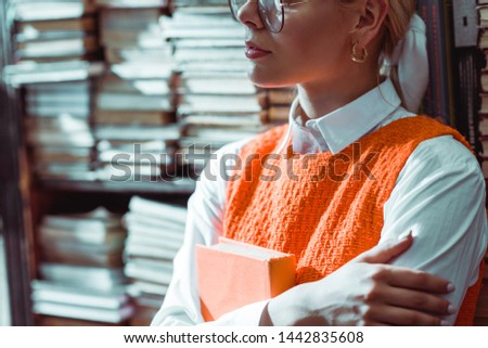 cropped view of woman in glasses holding orange book in library 