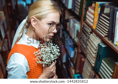 beautiful and blonde woman holding white flowers and looking away in library 