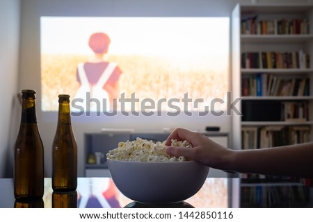 Home cinema entertainment: watching a film from a video projector in a room. Dim living room with a cinematic picture projected on the wall, human hand grabbing popcorn