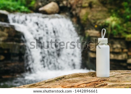 White bottle with water standing on a wooden trunk against the background of a river and a waterfall. The concept of pure natural drinking water