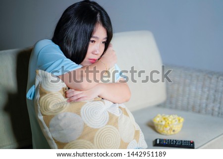 young beautiful and scared Asian Chinese teenager woman in fear watching horror scary movie at home sofa couch eating popcorn bowl holding remote controller in horrified face expression