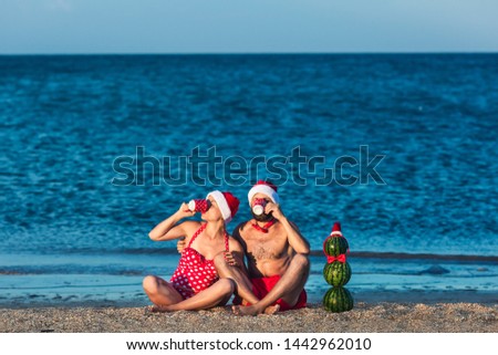 bearded man and girl in Santa's clothes are drinking from cups on seashore, and snowman made of watermelons is standing nearby. Summer Christmas Celebration