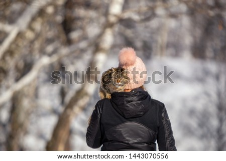 Winter picture. A tabby cat sits on the shoulder of a woman in a pink hat with a large pumpkin. A woman and a cat are walking together in the park. Sunny day. Pets. Cats.