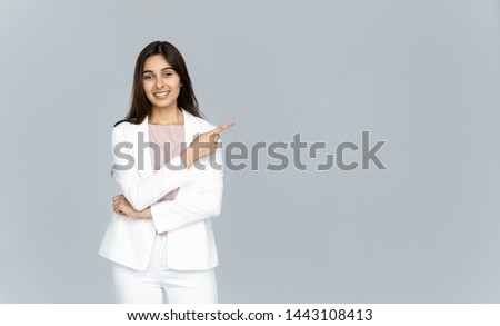 Happy indian young business woman wear white suit looking at camera pointing finger at copy space isolated on grey studio background, smiling beautiful confident hindu lady professional showing aside Royalty-Free Stock Photo #1443108413